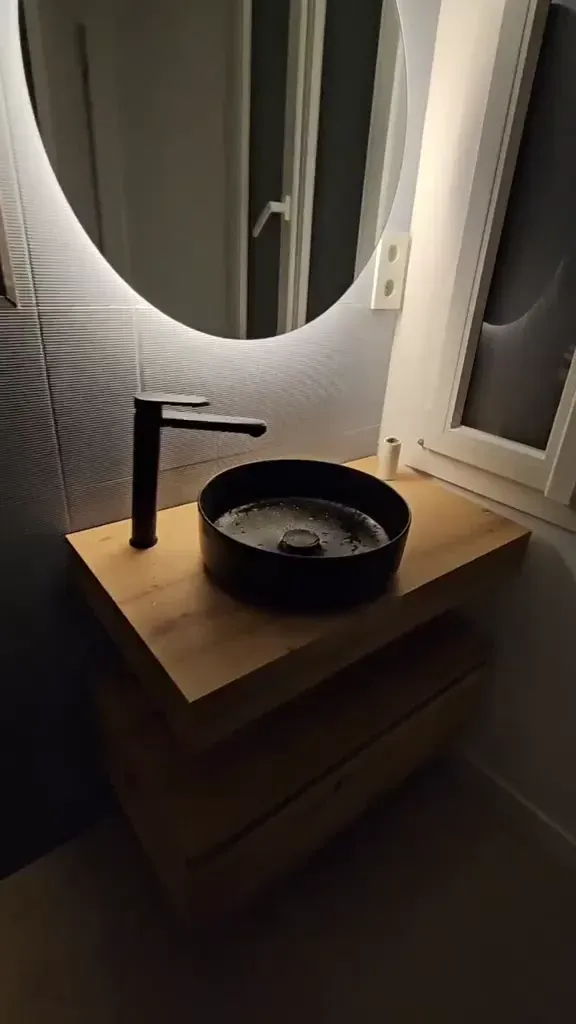 Lavabo negro sobre mueble de madera y espejo redondo con luz